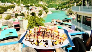 Une vue sur un paysage montagneux, avec une rivière, des bâtiments et une assiette de nourriture grillée au premier plan.
