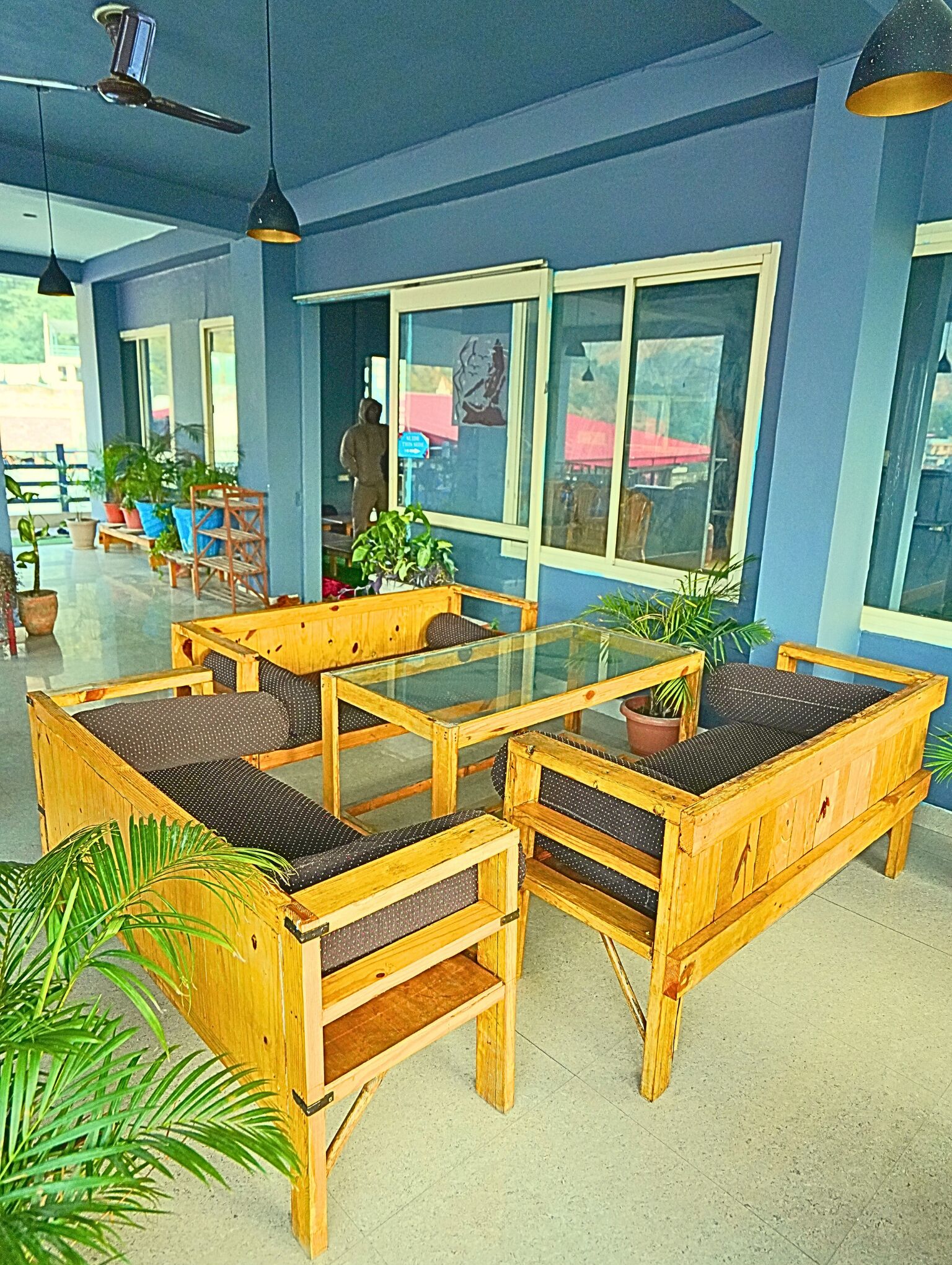 A seating area with wooden furniture and glass tables, surrounded by potted plants and large windows.