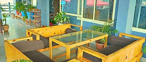 A seating area with wooden furniture and glass tables, surrounded by potted plants and large windows.