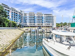 Een jachthaven met aangemeerde boten, een gebouw met meerdere verdiepingen op de achtergrond, en een heldere lucht.
