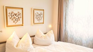 Un lit bien fait, avec une literie blanche et deux coussins décoratifs, une œuvre d’art encadrée représentant des cœurs accrochée au mur, et une fenêtre avec des rideaux.