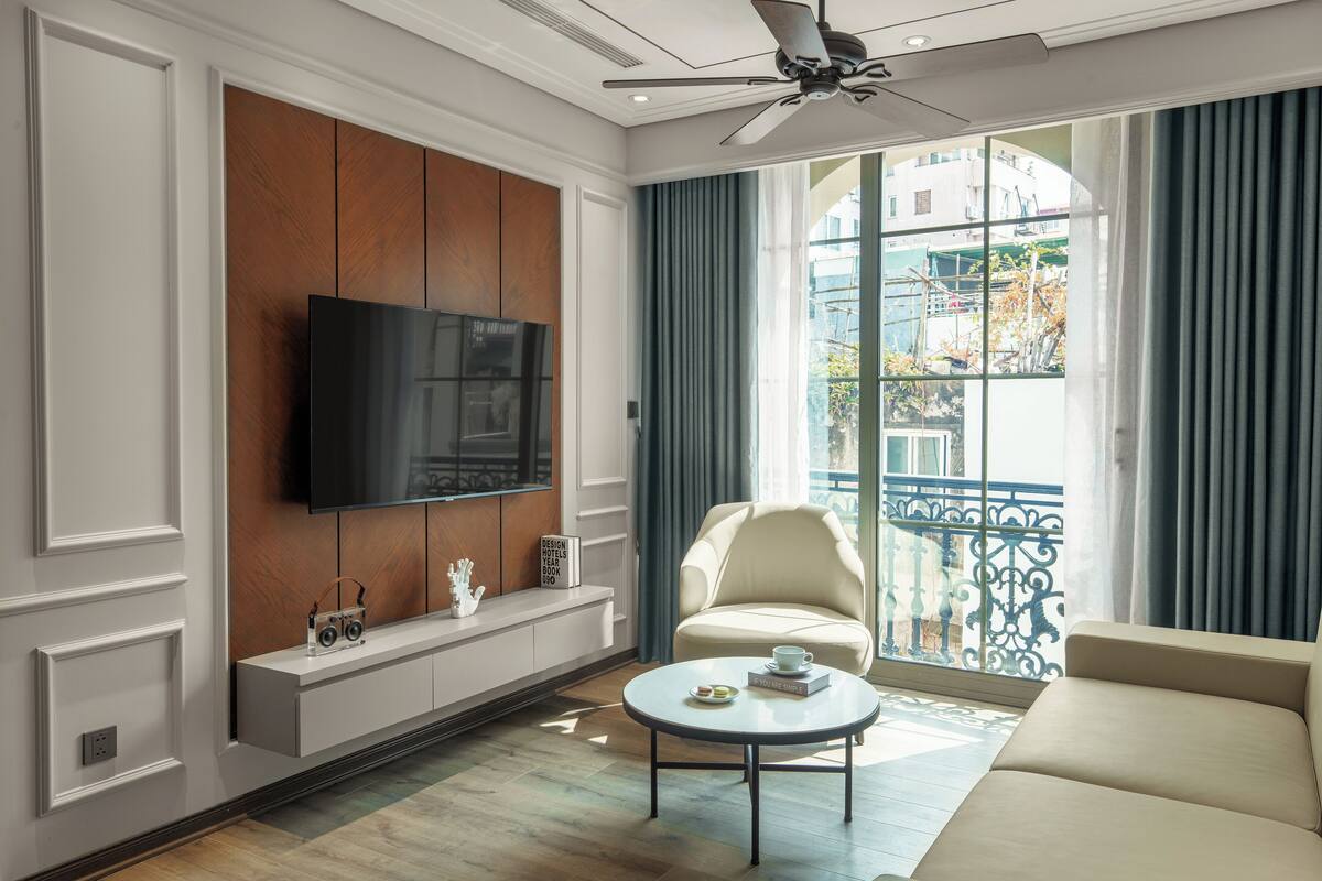 A modern living room with a flat-screen TV mounted on a wooden panel wall, a comfortable beige sofa, a round coffee table with a book and a cup, and a balcony with blue curtains and a view of buildings.