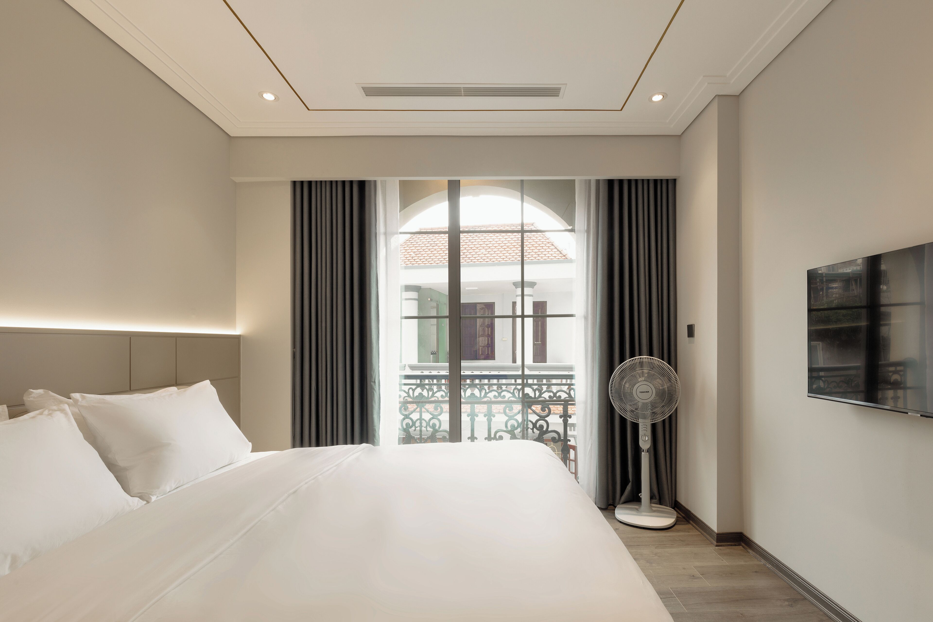 A modern hotel room with a large bed, a standing fan, a TV, and a balcony with a view.