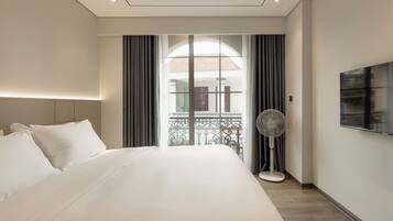A modern hotel room with a large bed, a standing fan, a TV, and a balcony with a view.