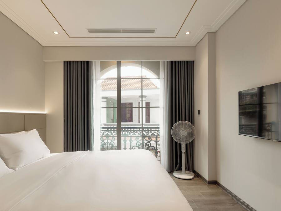 A modern hotel room with a large bed, a standing fan, a TV, and a balcony with a view.