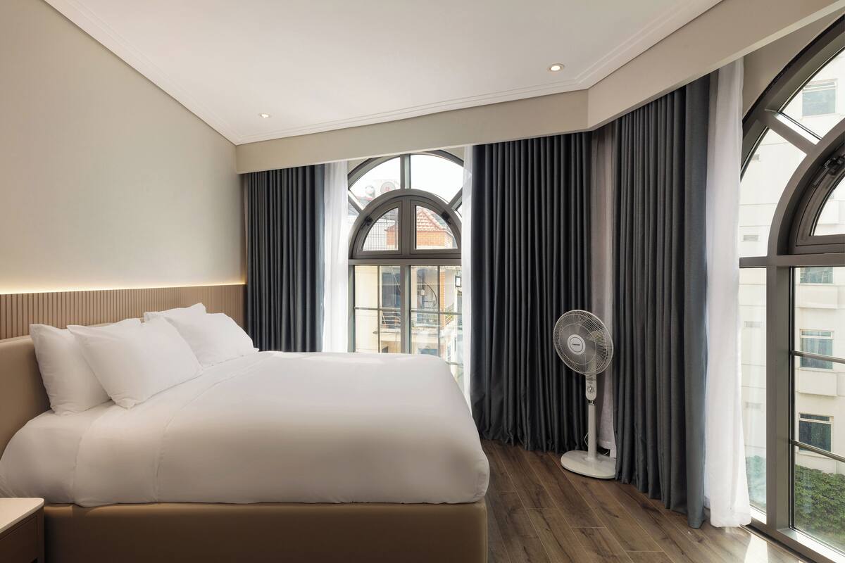 A hotel room with a large bed, a standing fan, and a view through a window with curtains.