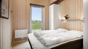 Een slaapkamer met houten wanden, een bed met wit beddengoed, een deur die uitkomt op een gazon en een plankje met een plant.