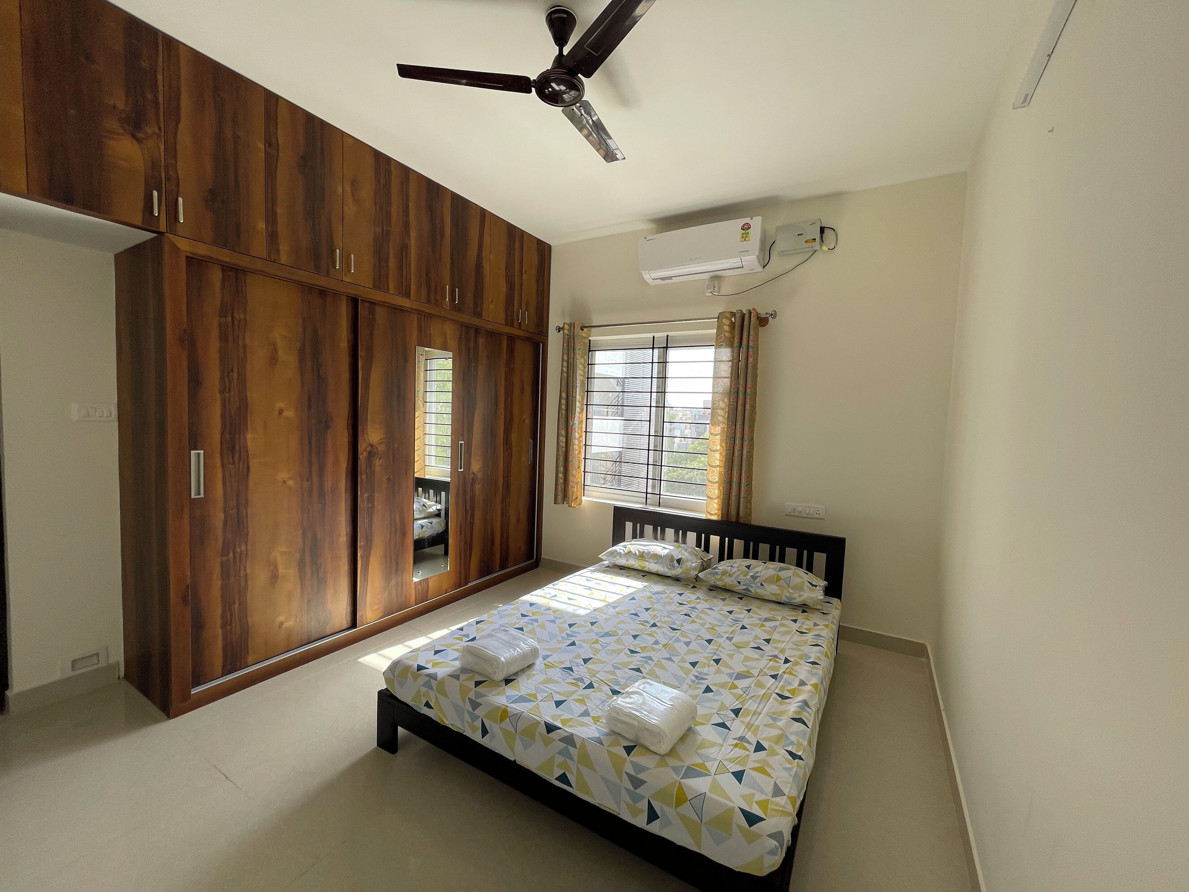 A bedroom with a bed, wardrobe, ceiling fan, air conditioning unit, and a window with curtains.