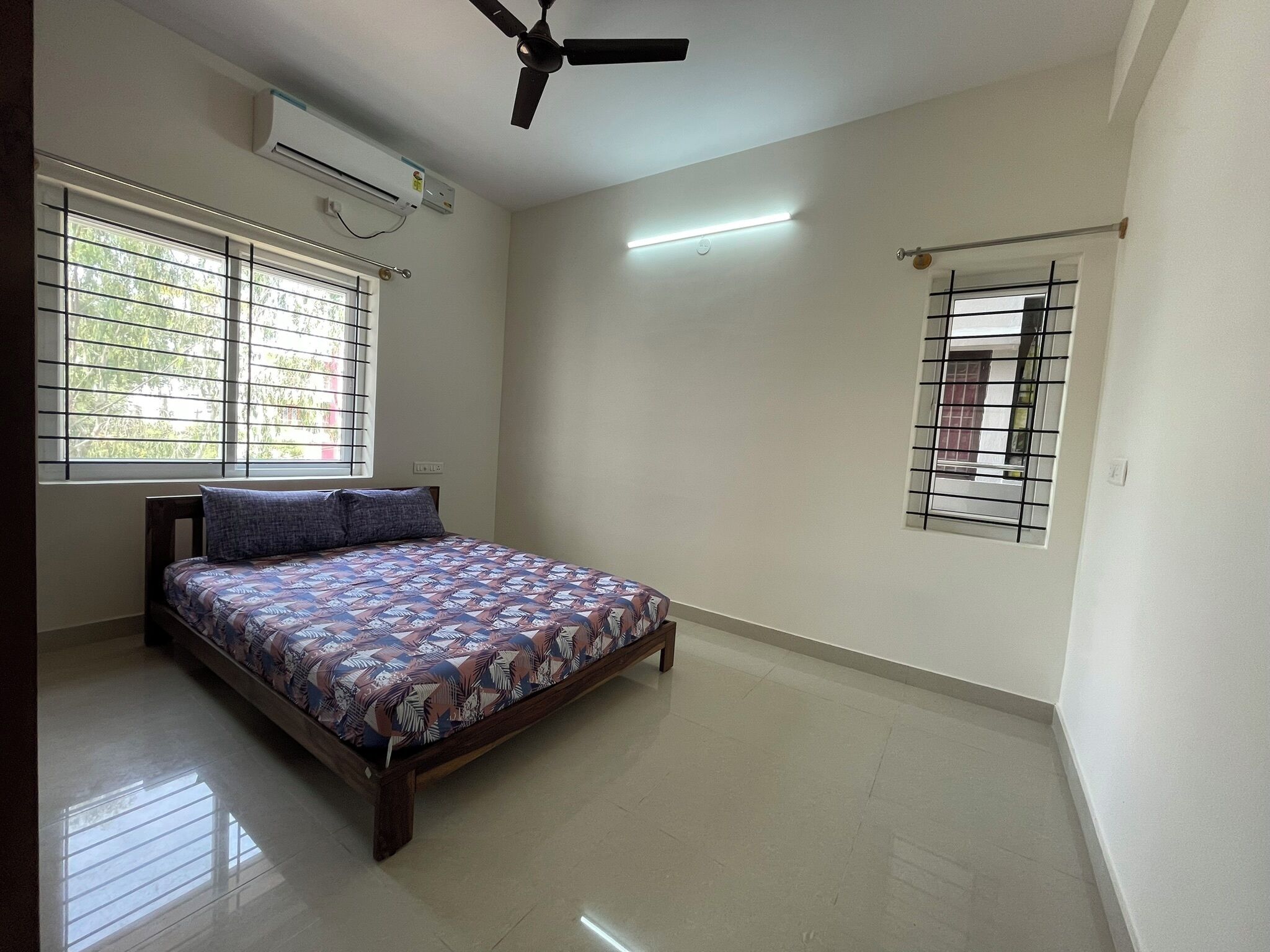 A bedroom with a bed, a ceiling fan, an air conditioning unit, a window with bars, and a small window with a metal grille.