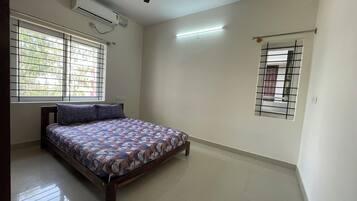 A bedroom with a bed, a ceiling fan, an air conditioning unit, a window with bars, and a small window with a metal grille.
