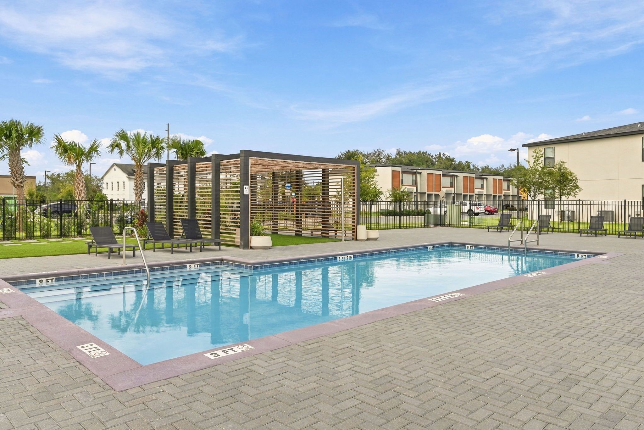 A swimming pool with a wooden deck and lounge chairs, surrounded by a fence and palm trees.