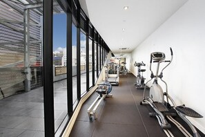 A gym with cardio machines and a bench, large windows, and a view of the city.