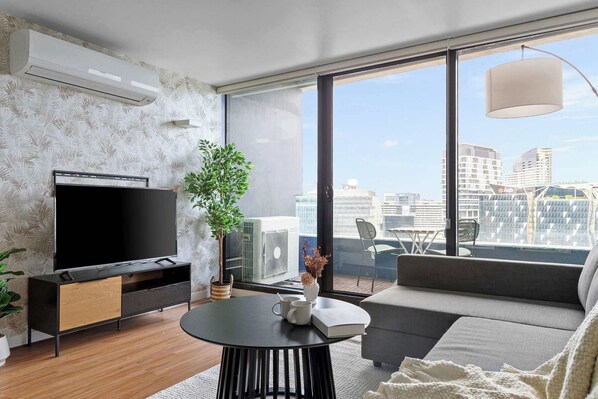 A modern living room with a flat-screen TV, a sofa, a dining table, and a large window offering a city view.