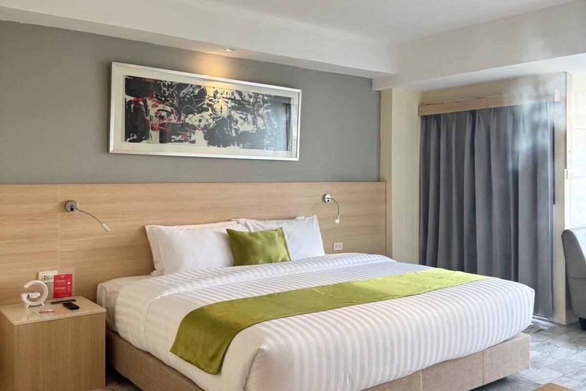 Une chambre d’hôtel moderne avec un grand lit, une table de chevet, une commode, un bureau, une chaise et une grande fenêtre avec des rideaux.