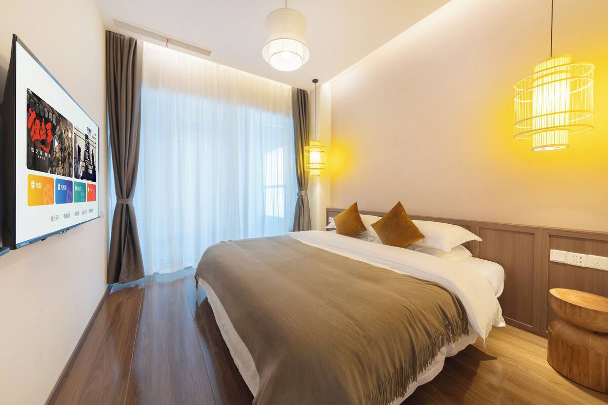 Ein Hotelzimmer mit einem großen Bett, einem Nachttisch aus Holz, einem an der Wand montierten Fernseher und einem Fenster mit Vorhängen.