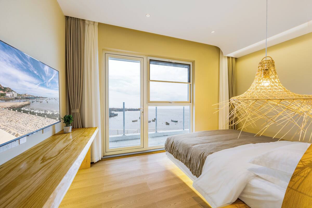 Ein Hotelzimmer mit großem Fenster, einem Bett, einer Holzbank und einer Pendelleuchte.