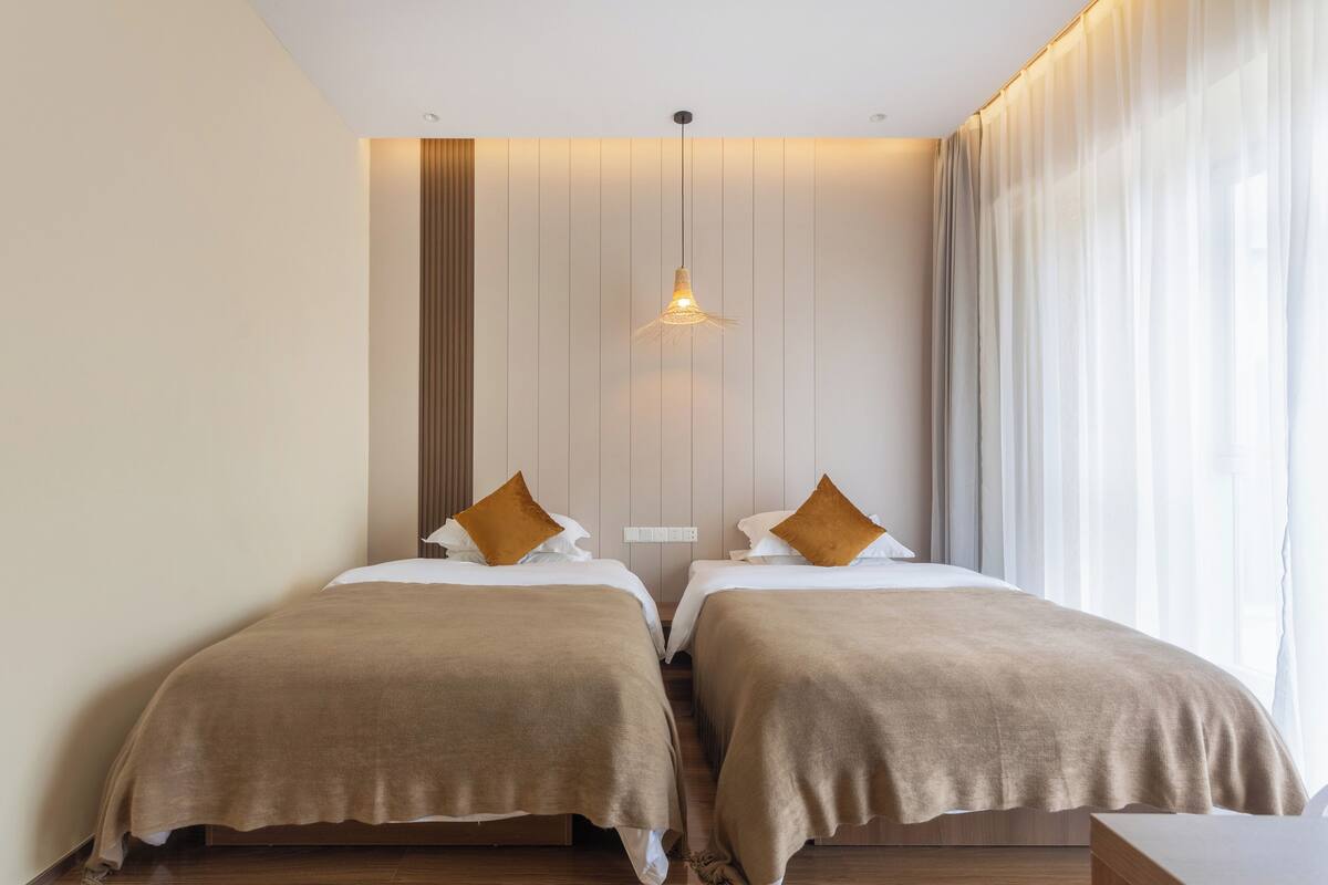 Ein Hotelzimmer mit zwei Betten, einer Hängelampe, einer Wand aus Holzpaneelen und einem großen Fenster mit Vorhängen.
