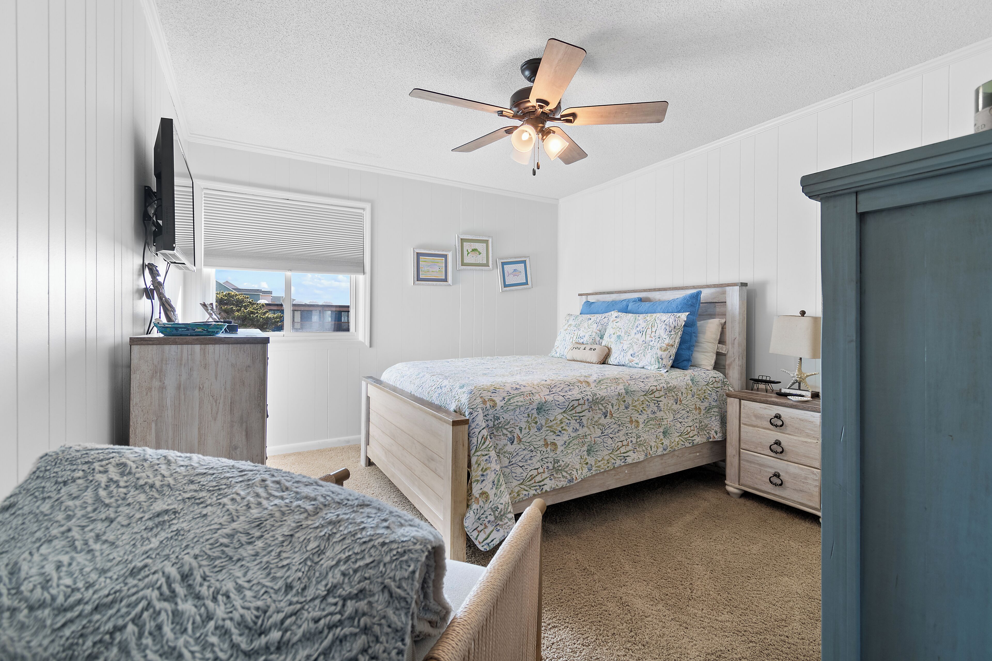 A bedroom with a bed, a dresser, a nightstand, a ceiling fan, a window, and a wall-mounted TV.