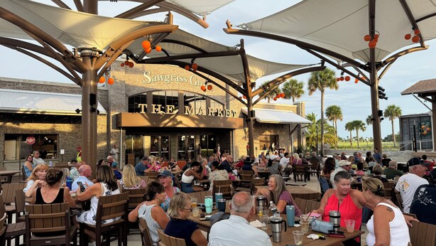 Et udendørs spiseområde med træborde og -stole, en murbygning med skiltet "Sawgrass Grill THE MARKET", og en stor markise over hovedet.
