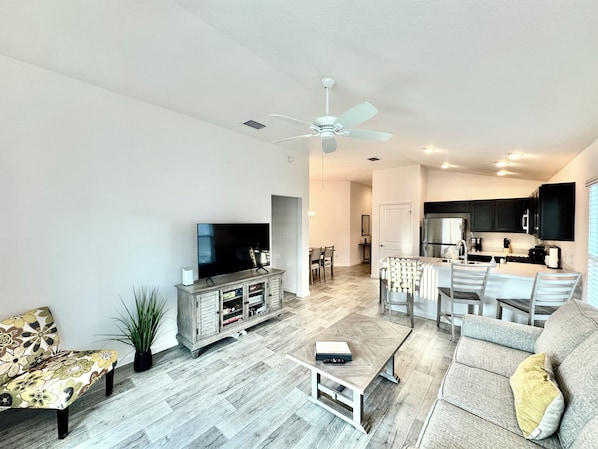 A modern living room with a flat-screen TV, a coffee table, a sofa, and a dining area with chairs.