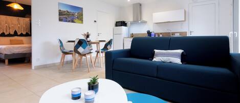 Een moderne woonkamer met een blauwe bank, een ronde witte salontafel, en een eethoek met twee stoelen en een kleine tafel.