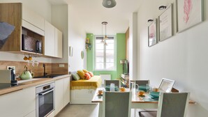 Een kleine keuken met een ingebouwde oven, een magnetron en een koffieapparaat. Er is een eethoek met een tafel en stoelen, en een raam dat voor natuurlijk licht zorgt.