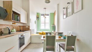 Een kleine keuken met witte kastjes, een magnetron en een spoelbak. Er is een eettafel met stoelen en een raam met groene luiken.
