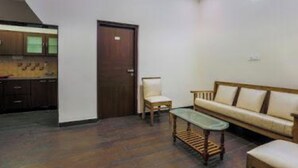 A living room with a sofa, a small table, and a chair. There is a wooden door and a kitchen area visible in the background. - SRJ Thaneesh Residency (Ootacamund)