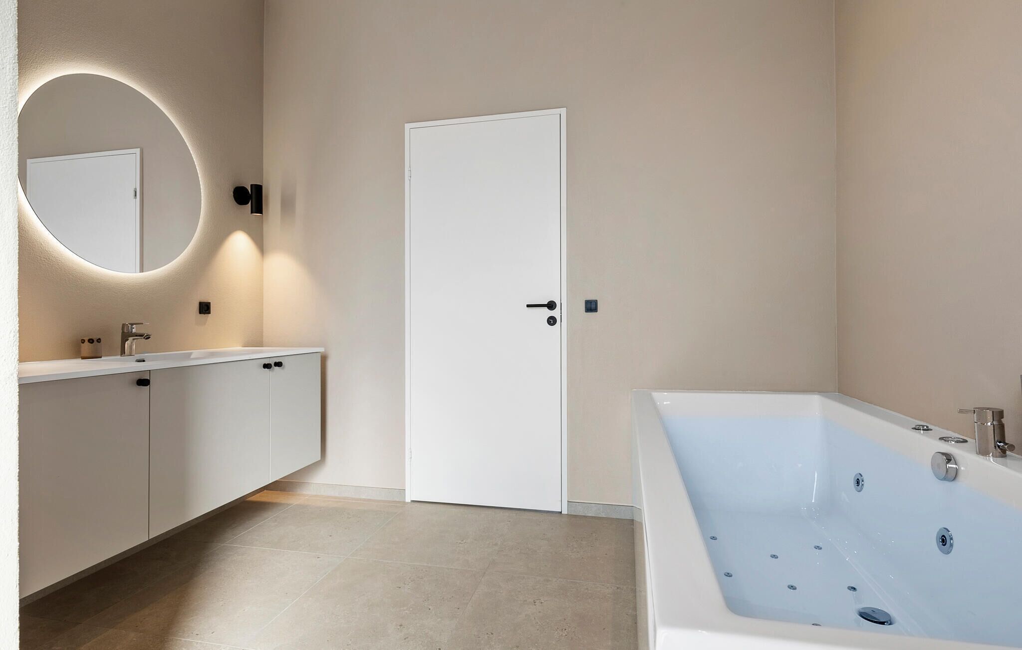A modern bathroom with a large bathtub, a round mirror, and a white door.