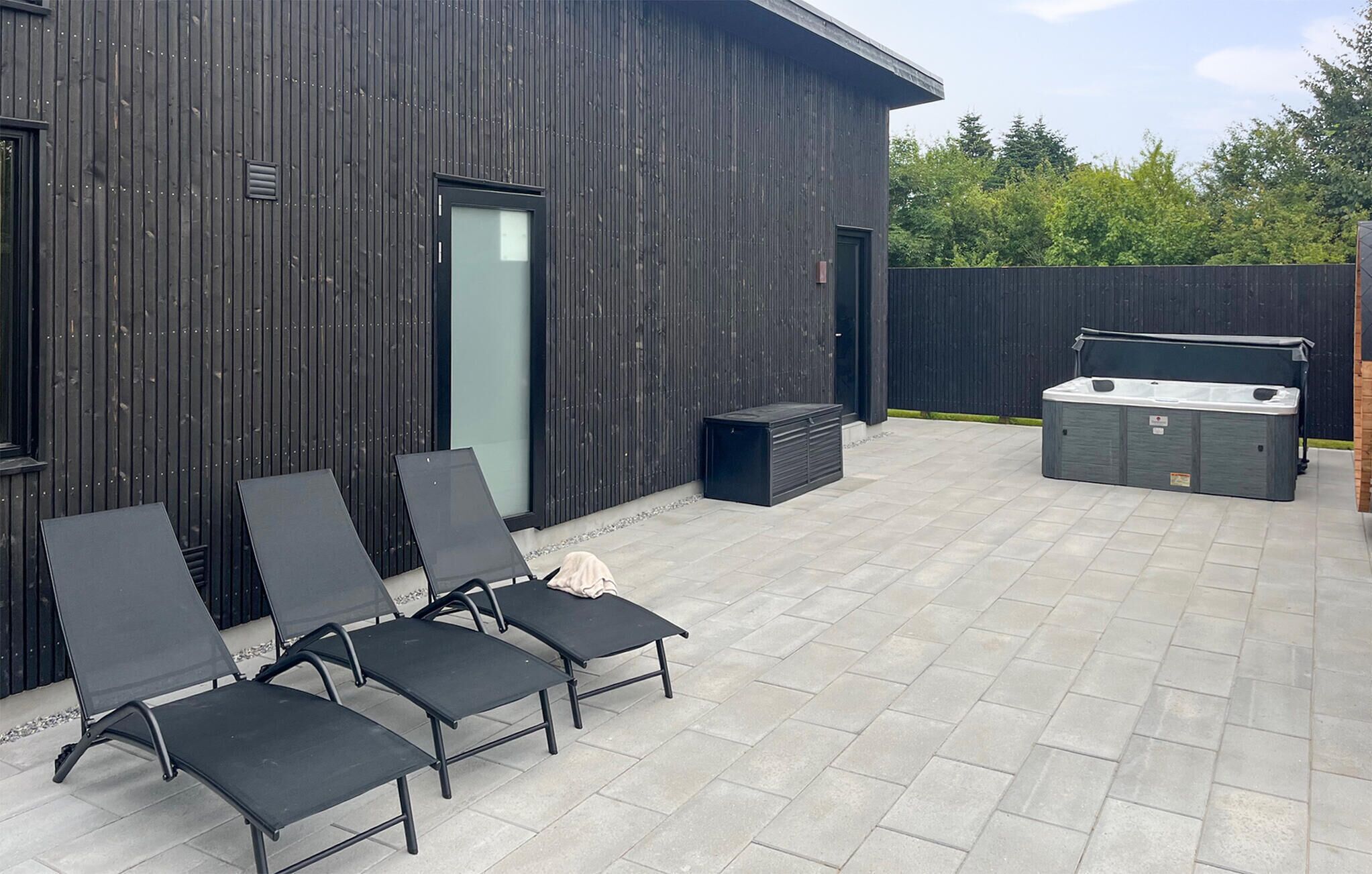 Une terrasse extérieure avec quatre chaises longues noires, un jacuzzi et un bâtiment en bois doté d’une porte en verre.
