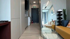 A modern reception area with a glass coffee table, a sofa with cushions, a washing machine, and a flat-screen TV mounted on the wall. - The Bayment Residence (Busan)