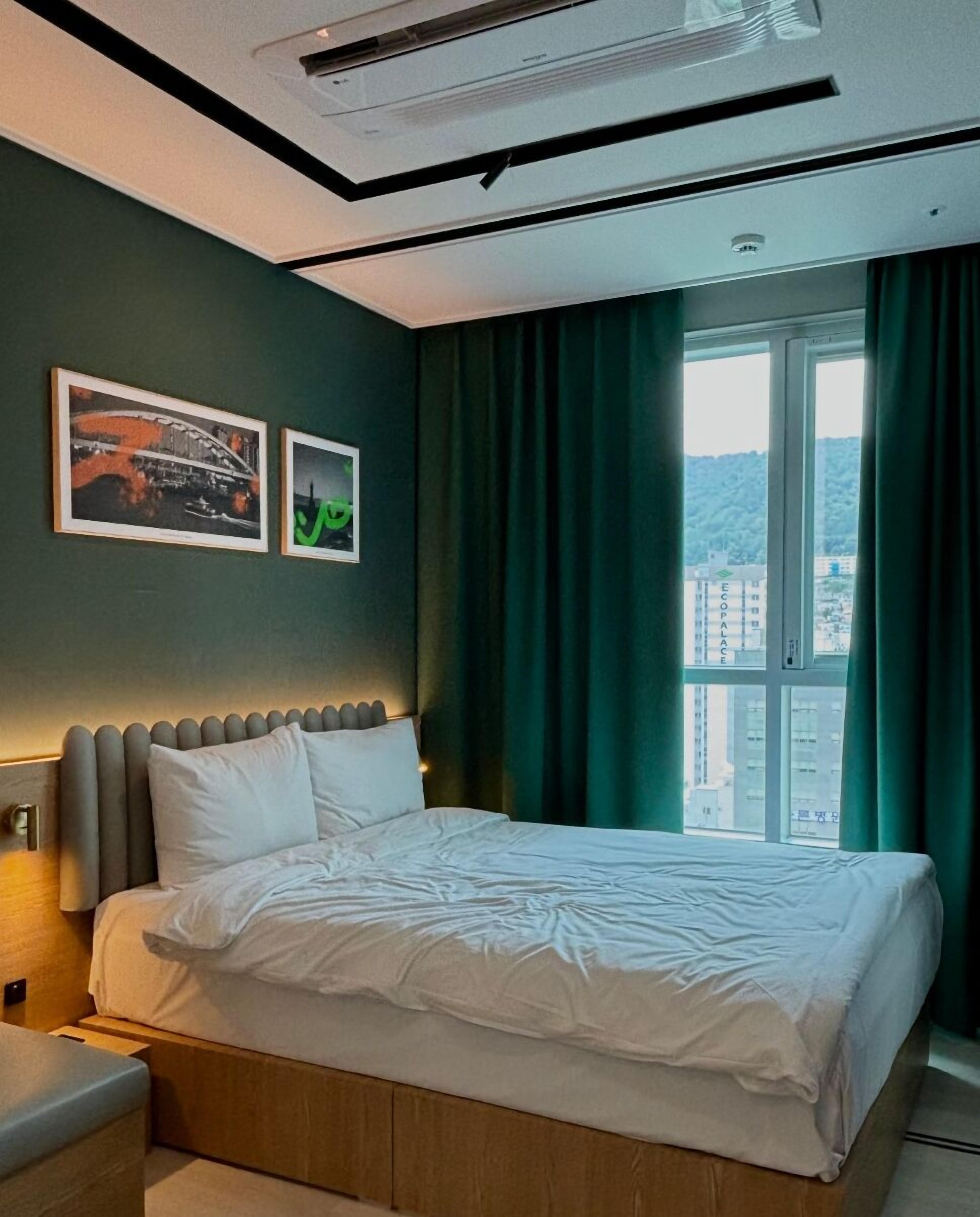 Una habitación de hotel moderna con una cama grande, una mesita de noche, un cabecero con iluminación integrada y una ventana con cortinas.
