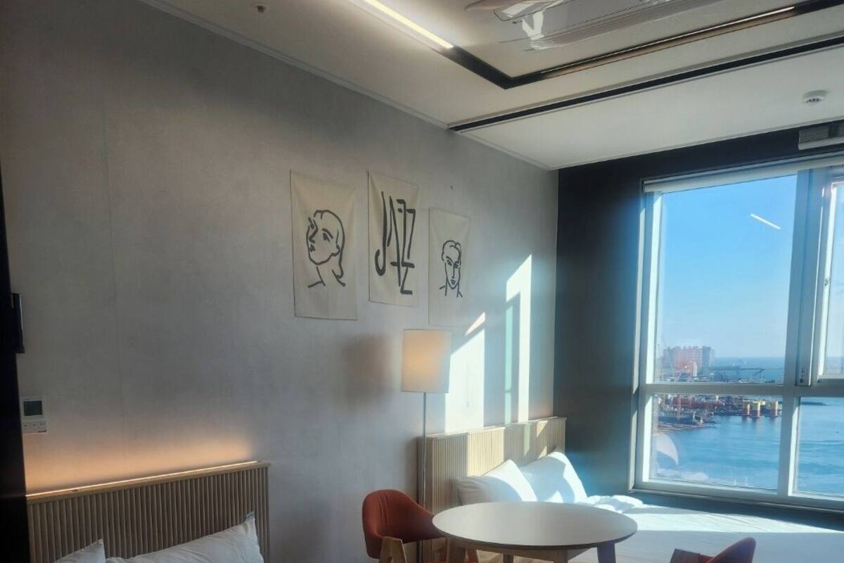 A hotel room with a large bed, a round table with two chairs, a window with a view of the sea, and wall art.
