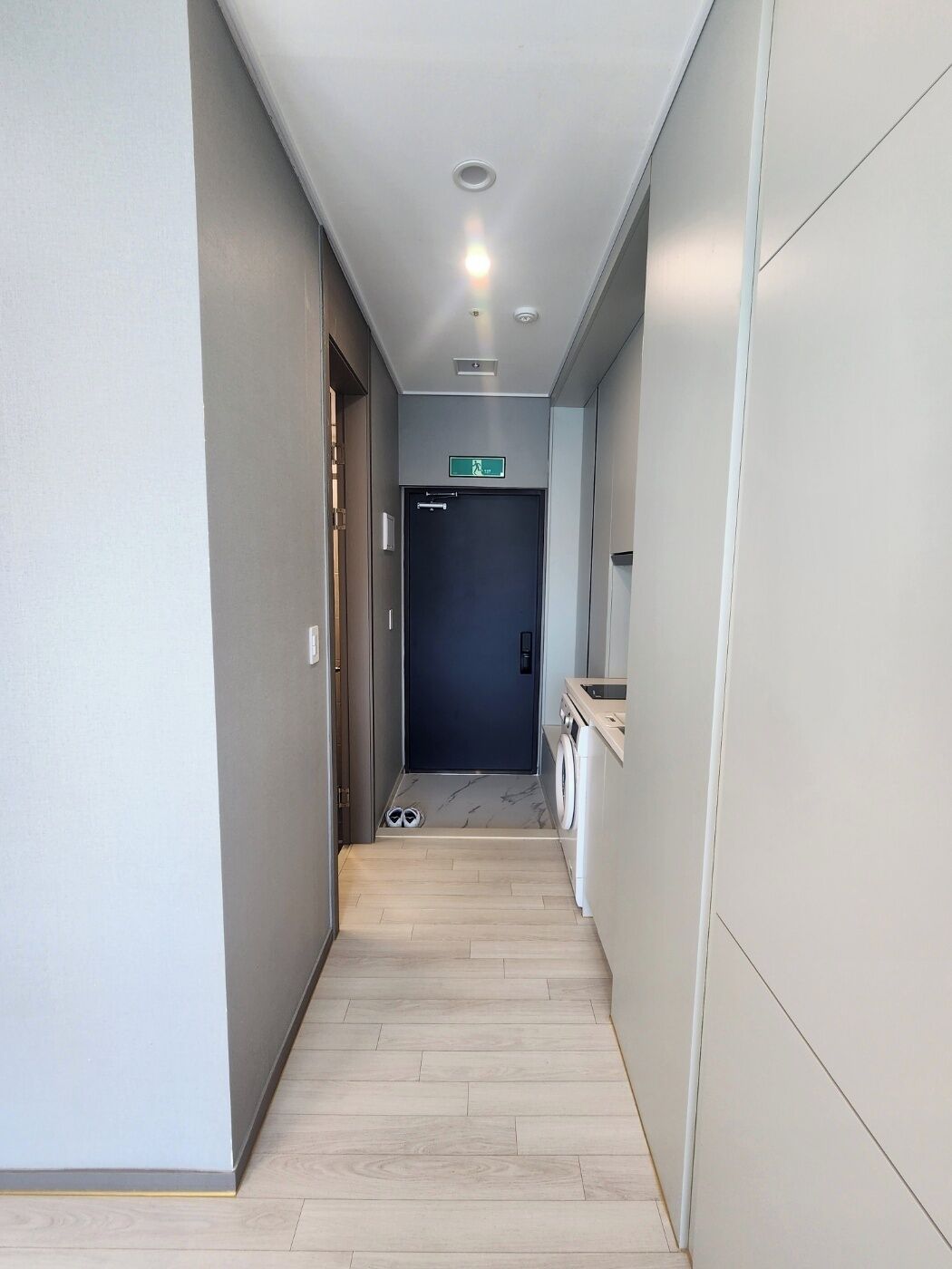 Un couloir étroit, avec une porte sombre au fond, une petite marche et une paire de chaussures posées sur le sol.