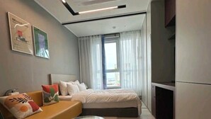A modern bedroom with a bed, a glass coffee table, a sofa with cushions, and a view of the outdoors through a window. - The Bayment Residence (Busan)