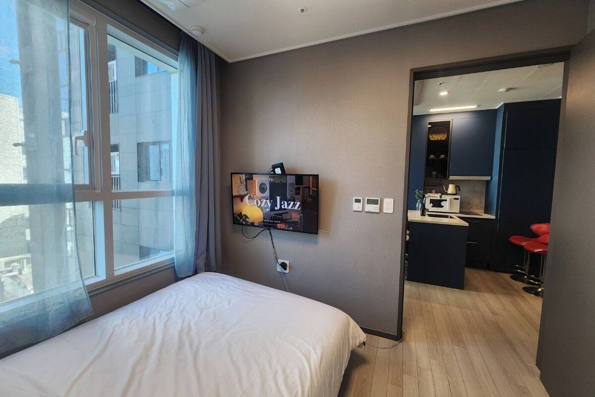A modern hotel room with a large bed, a TV displaying 'Cozy Jazz', a kitchenette, and a bathroom visible through an open door.