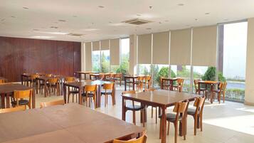 Un comedor amplio con varias mesas y sillas de madera, ventanas grandes que ofrecen vista a zonas verdes y un techo moderno con iluminación empotrada.