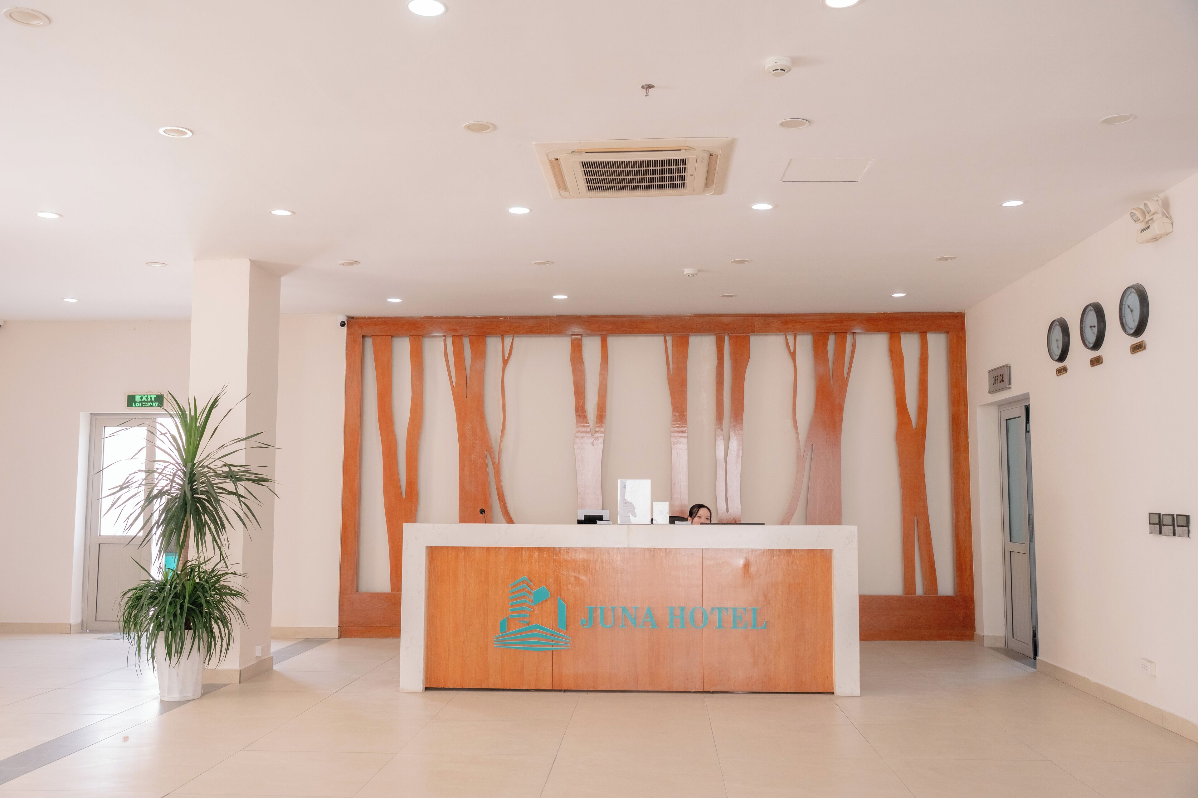 A reception area with a wooden reception desk labeled "JUNAHOTEL," a potted plant, and a wall-mounted clock.