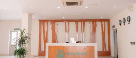 A reception area with a wooden reception desk labeled "JUNAHOTEL," a potted plant, and a wall-mounted clock.