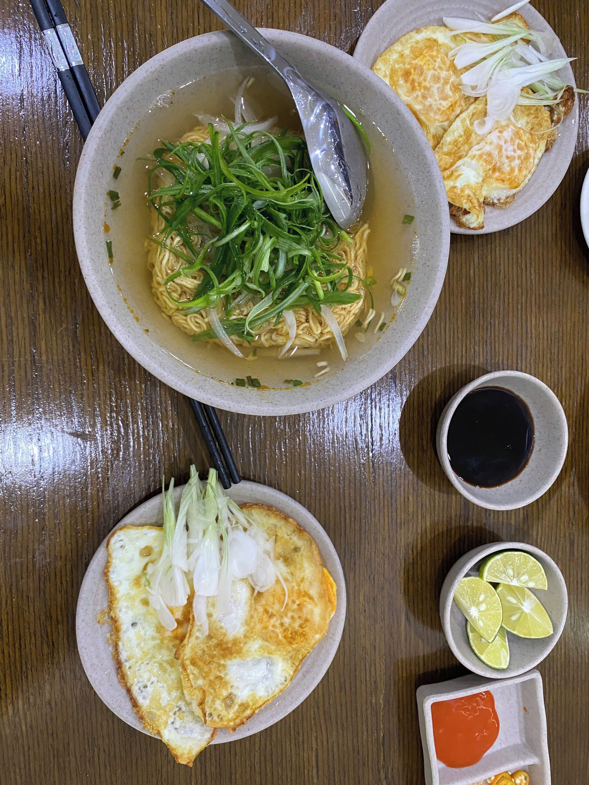 Un tazón de ramen con cebolletas, un plato de huevo frito y dos recipientes pequeños con salsa sobre una mesa de madera.