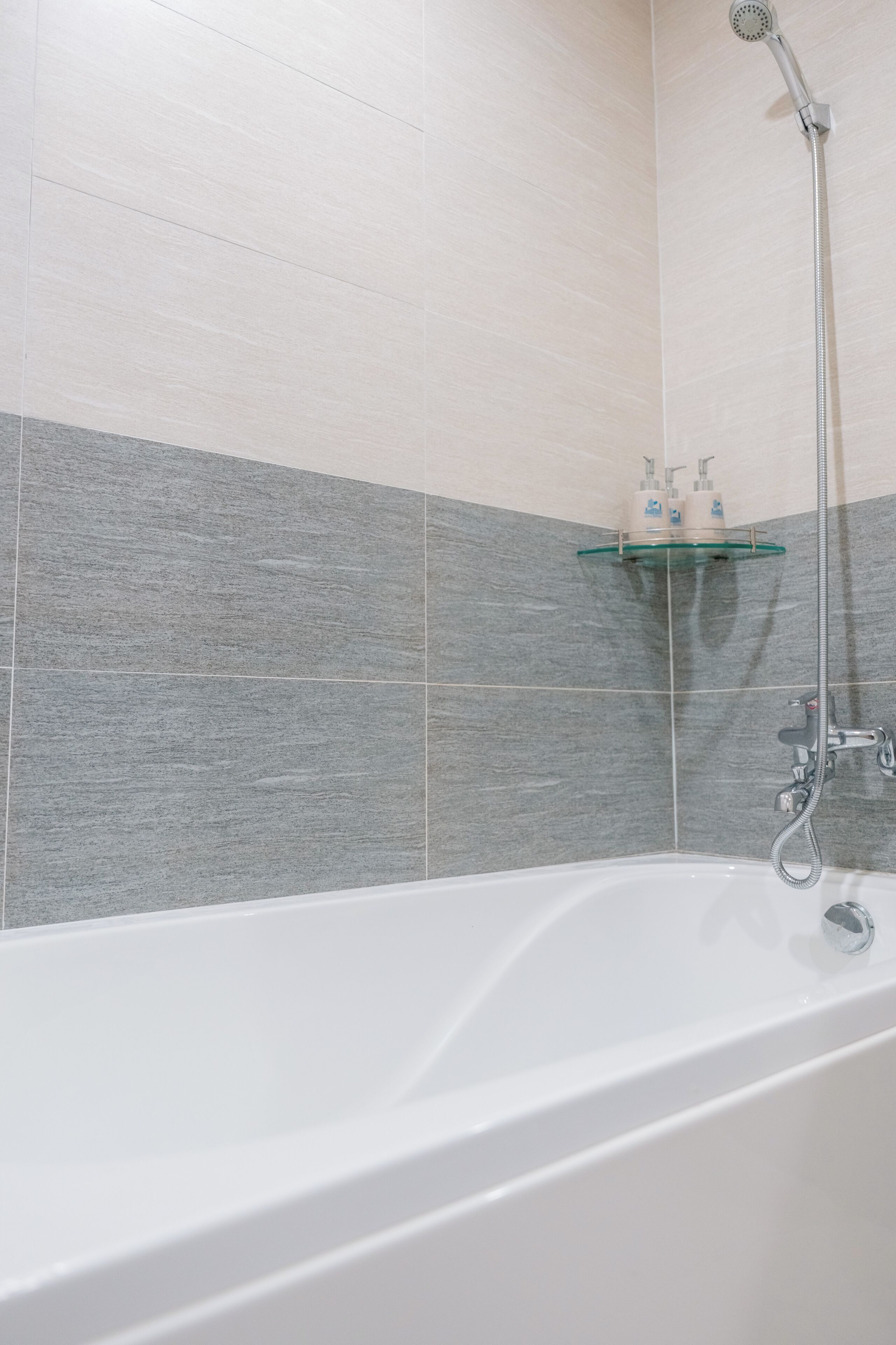 Un baño moderno con una bañera blanca, un estante de vidrio con artículos de aseo, y paredes con azulejos grises y blancos.