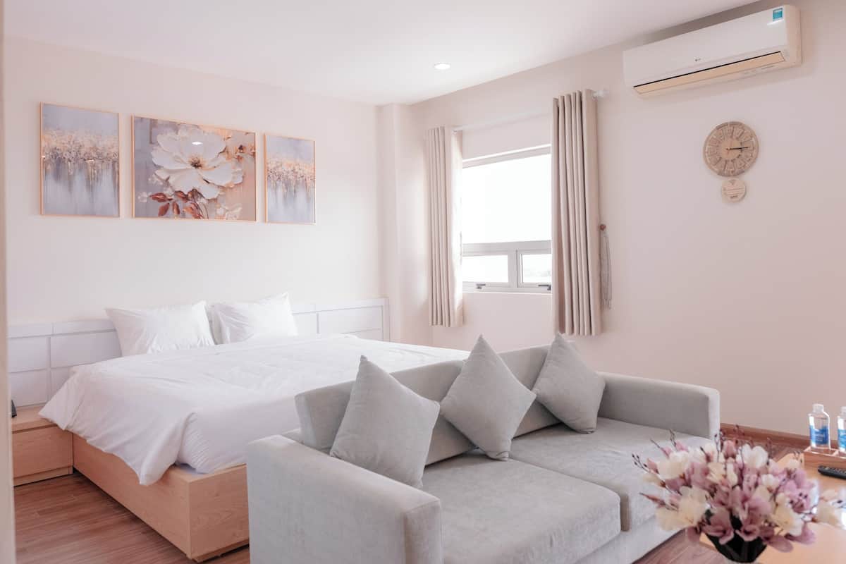 Una camera da letto moderna con un letto, un divano grigio e una finestra con tende. Sono presenti condizionatori fissati al muro e una sveglia.