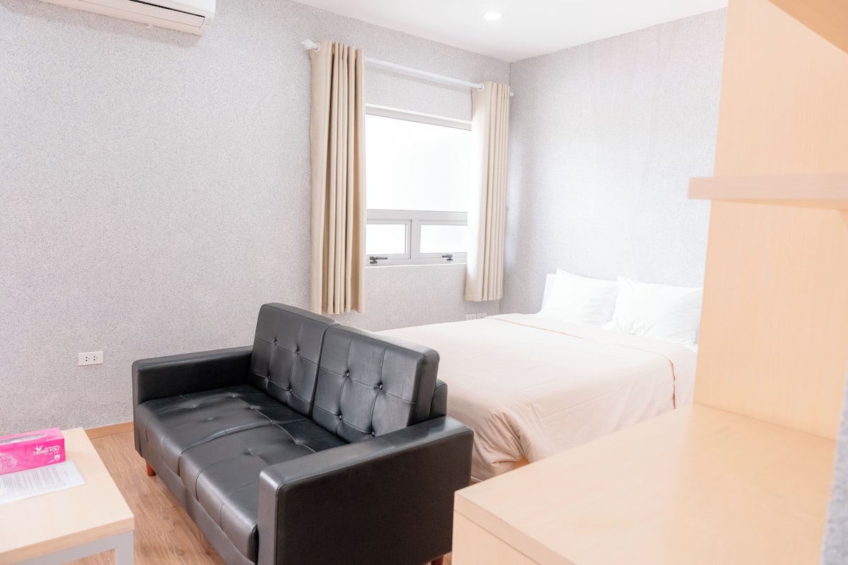 Una camera d'albergo con un divano in pelle nera, un letto con biancheria bianca, una finestra con tende beige e un tavolino con una scatola rosa.