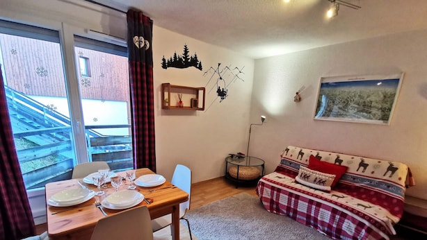 Une pièce chaleureuse avec une table à manger dressée pour deux personnes, un canapé recouvert d’un tissu à motifs, une étagère murale agrémentée d’objets décoratifs et un tableau encadré accroché au mur.