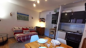 Un espace de vie compact comprenant un coin cuisine, un coin repas aménagé pour quatre personnes et un canapé confortable agrémenté d’un plaid à motifs.