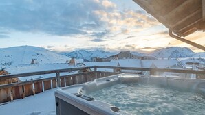 Ein Whirlpool mit Holzterrasse, umgeben von schneebedeckten Bergen und blauem Himmel.