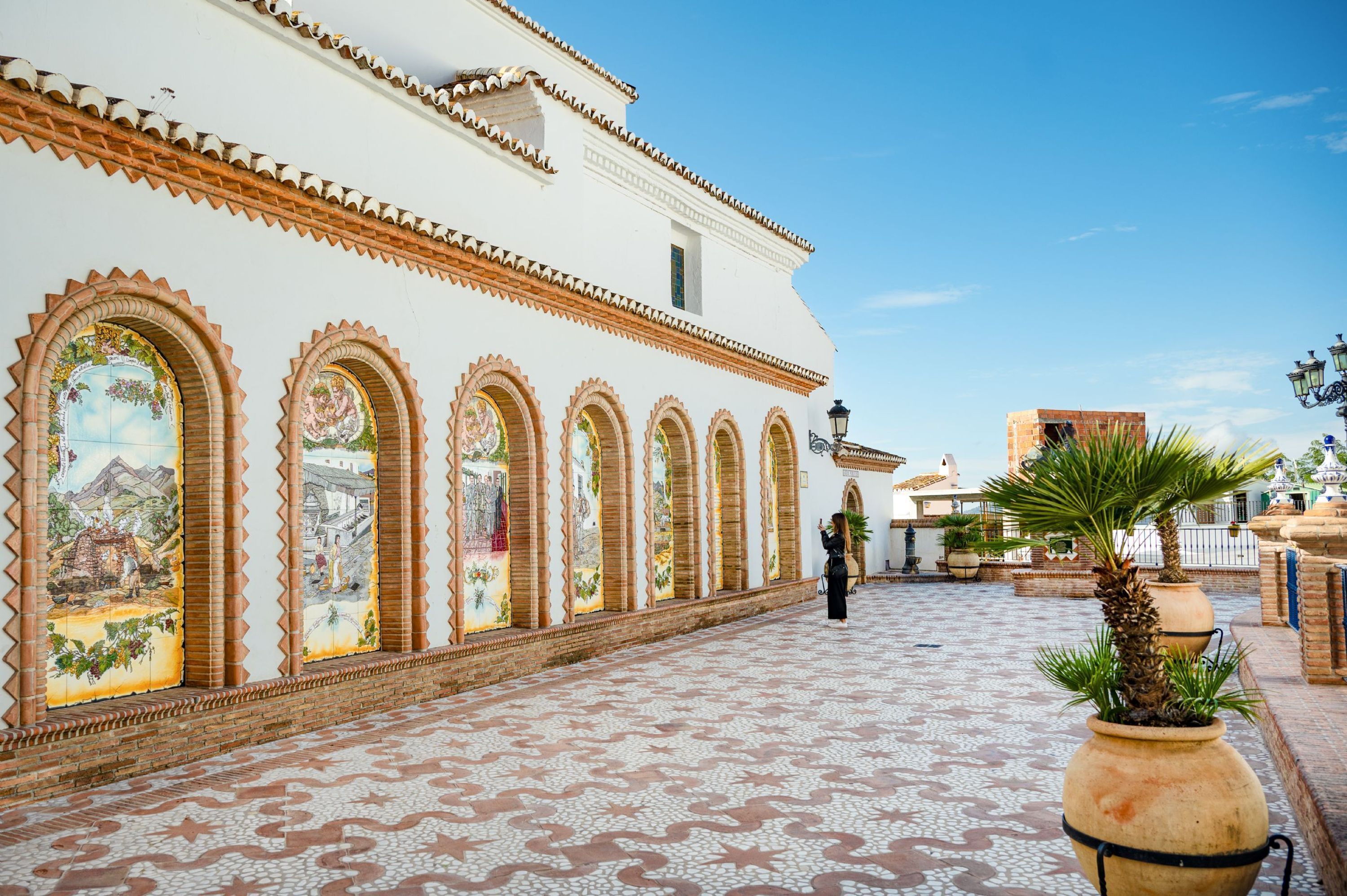 A courtyard with a tiled floor, white building with arched windows, and a person walking.
