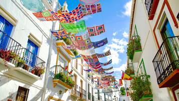 A narrow street with white buildings, balconies with plants, and colorful hanging decorations.