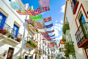 Una calle estrecha con edificios blancos, balcones con plantas y decoraciones colgantes coloridas.