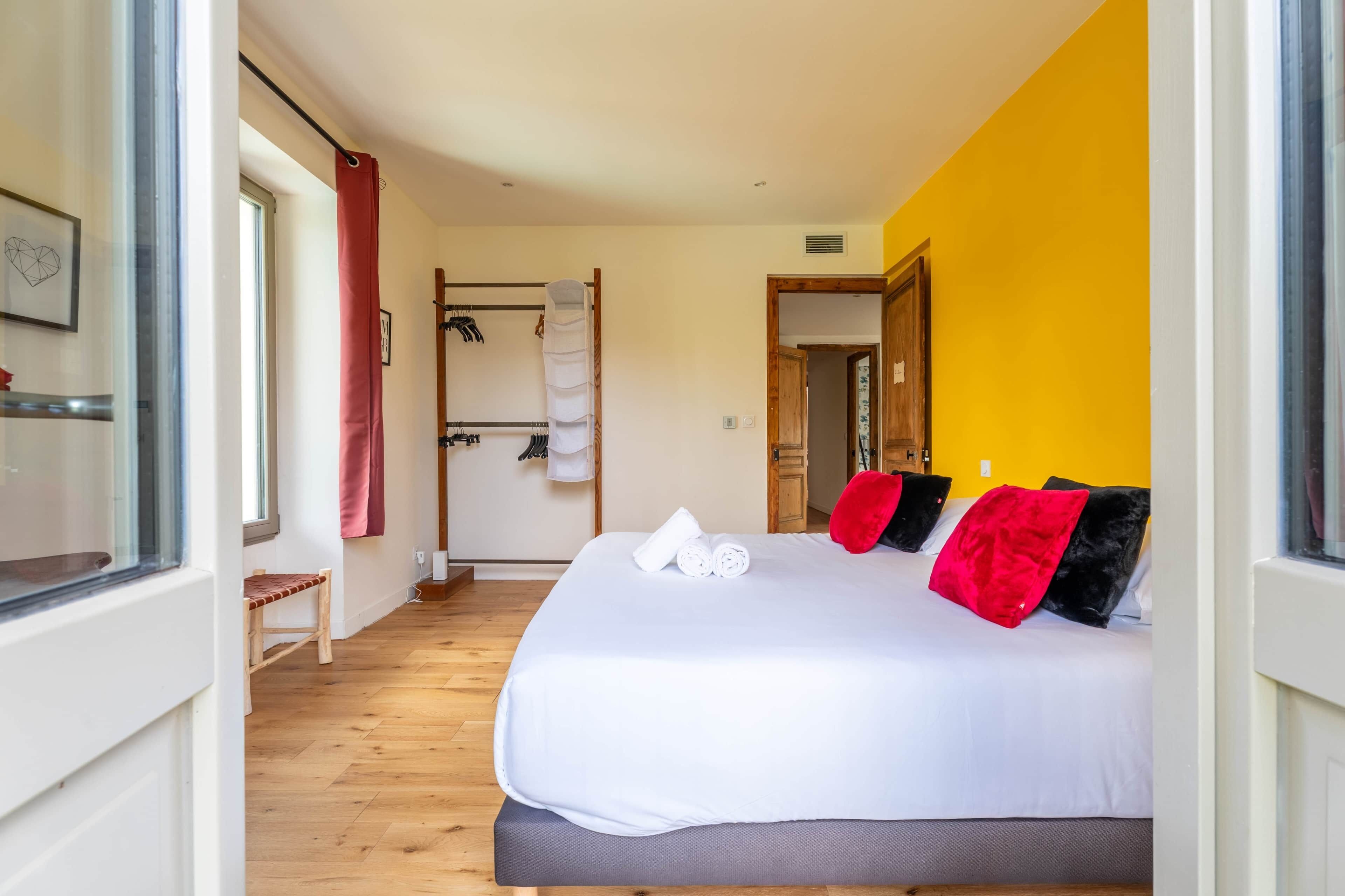 A hotel room with a bed, a wooden floor, a yellow wall, and a door leading to another room.